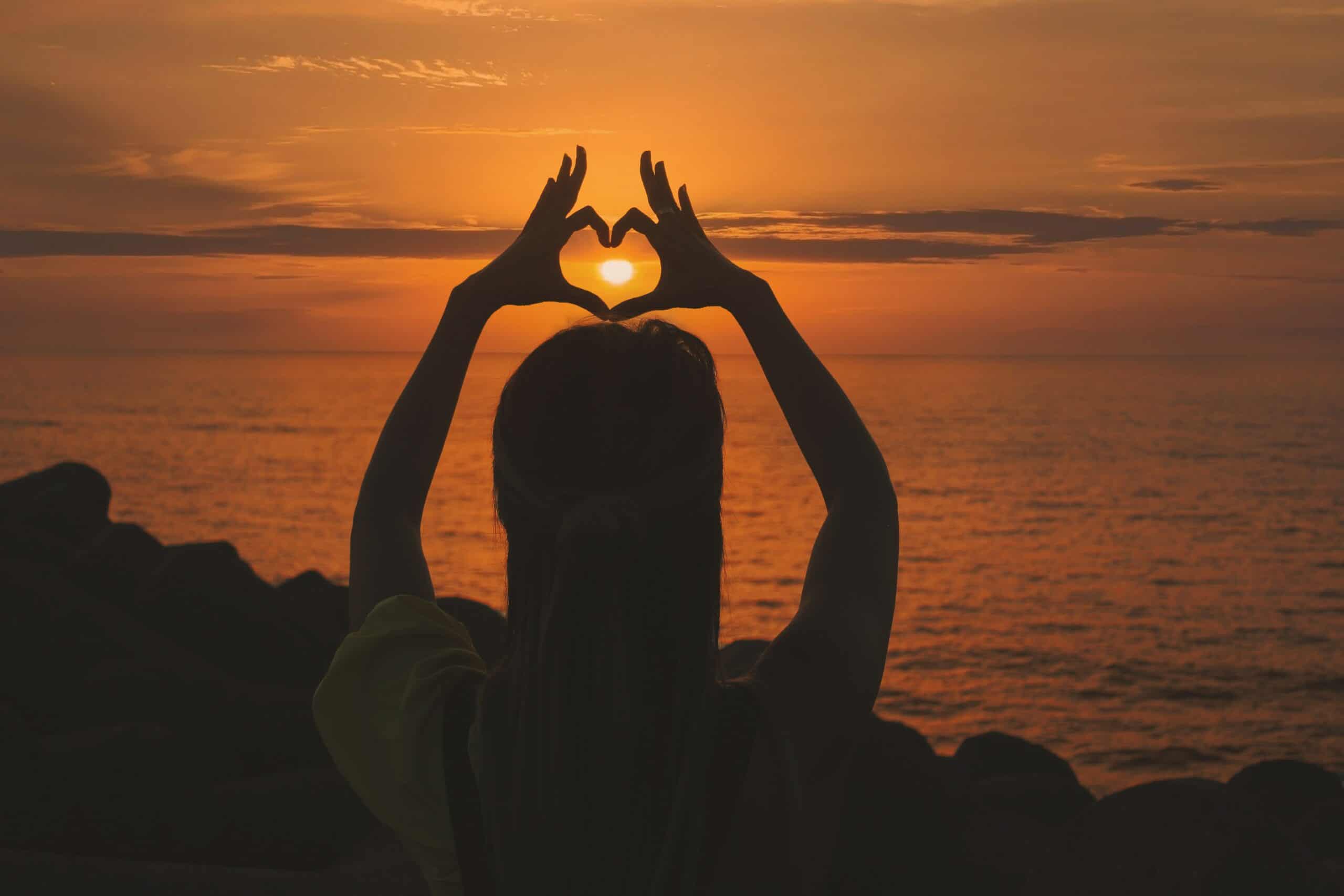 A silhouette of the back of a person standing next to the ocean with an orange sunset and they are holding their arms above their head with hands forming a heart shape and the sun is in the middle
