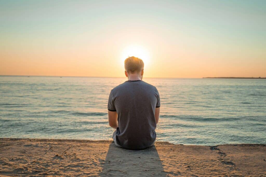 The back of a man sitting alone on a wall at the edge of the ocean at sunset