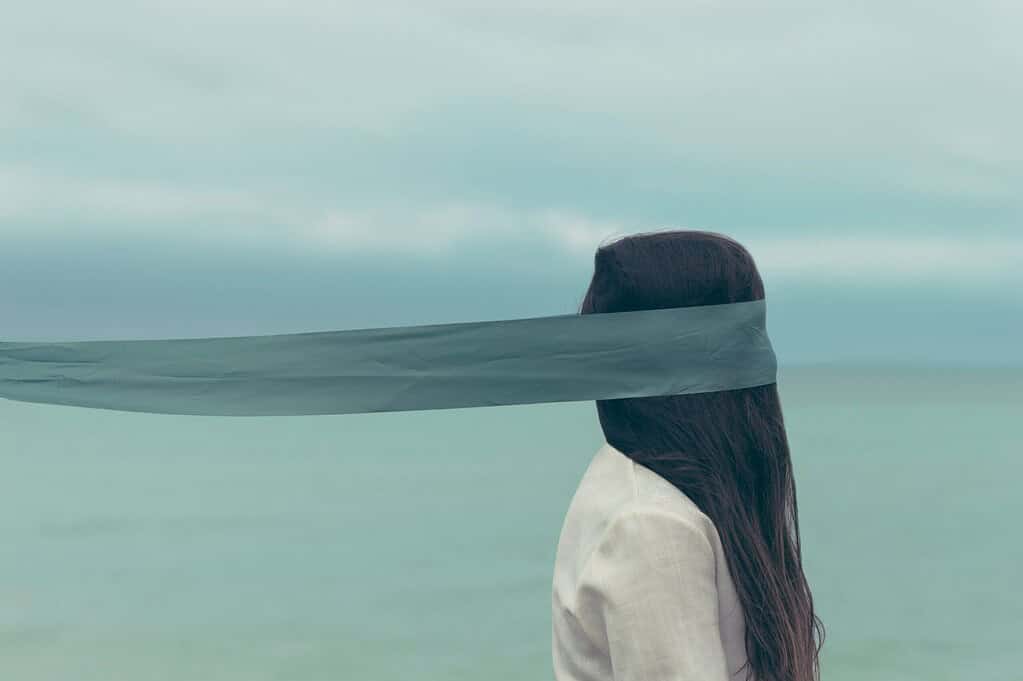 Image of a girl in front of the sea, who has a very long scarf blindfold covering her face and pulling her