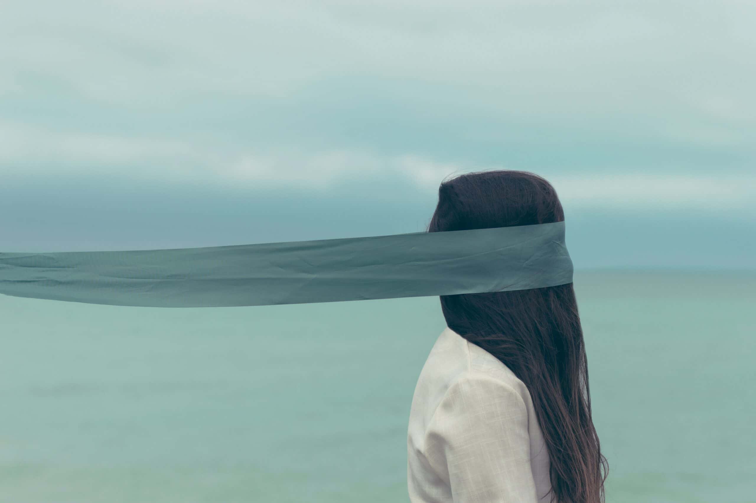 Image of a girl in front of the sea, who has a very long scarf blindfold covering her face and pulling her